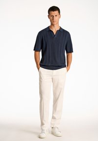 Laivastonsininen lyhythihainen polo, jossa on uritettu pinta, kolmen napin napitus, yhdessä kermaisten housujen ja valkoisten tennareiden kanssa.