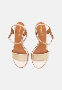 GEOX sandals with gold metallic straps, brown perforated insoles, flat sole, and buckle closure. Smooth texture and minimalistic design.