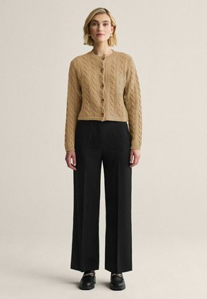 Nainen pukeutuneena vaaleanruskeaan kaapelikuvioiseen napilliseen neuletakkiin, mustiin leveälahkeisiin housuihin ja mustiin loafer-kenkiin, joissa on kultainen ketjupainatus, seisomassa yksiväristä taustaa vasten.