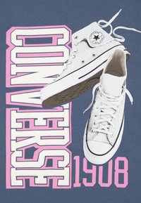 Bílé plátěné vysoké tenisky Converse se zaobleným černým logem, umístěné na modrém pozadí s velkým růžovým a bílým textem "CONVERSE 1908".