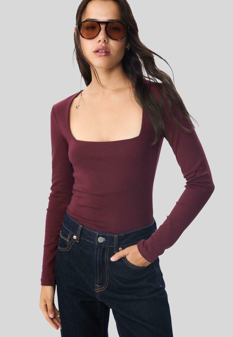 Burgunderfarbener Jumpsuit mit langen Ärmeln und quadratischem Ausschnitt, kombiniert mit dunkelblauen, hochtaillierten Jeans aus Denim mit goldfarbenen Nähten und einem vorderen Knopfverschluss.