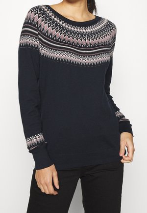 Kobieta ubrana w granatowy sweter z różowo-białymi wzorami diamentowymi na klatce piersiowej i mankietach, w połączeniu z czarnymi spodniami.