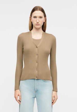 Kvinde iført en tætsiddende beige ribstrikket cardigan med knapper over en matchende top og lyseblå højtaljede jeans mod en ensfarvet baggrund.