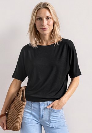 Blondynka ubrana w czarną bluzkę z krótkim rękawem i jasnoniebieskie jeansy, trzyma plecioną brązową torebkę, jedną ręką w kieszeni.