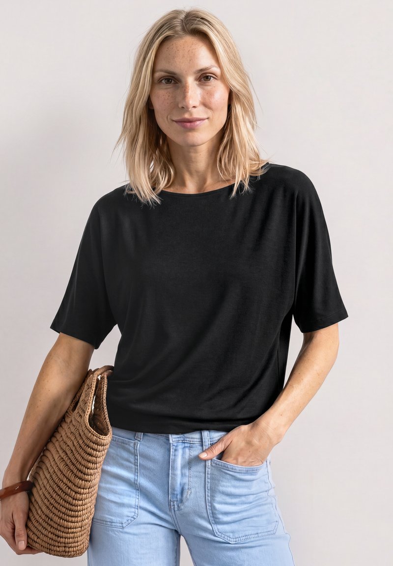 Femme blonde portant un haut noir à manches courtes et un jean bleu clair, tenant un sac à main marron tressé avec une main dans la poche.