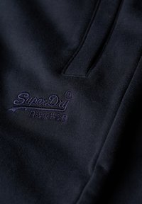 Tissu foncé avec logo SuperDry brodé et petits caractères japonais en dessous, montrant la texture et les détails de couture.