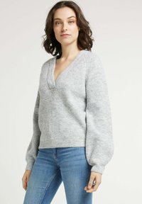 Pull en maille grise avec un col en V, de longues manches bouffantes et un motif texturé. Porté avec un jean bleu clair. Matériau doux et confortable.