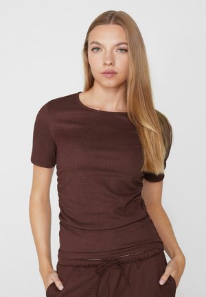 Junge Frau mit langen blonden Haaren, die ein enganliegendes braunes Kurzarmoberteil und passende Hose trägt, mit den Händen in den Taschen vor weißem Hintergrund.