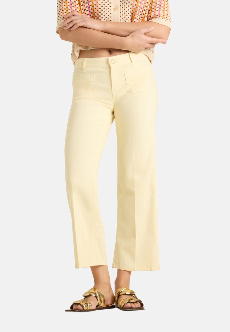 Person trägt blassgelbe kurze Hosen, mehrfarbiges gehäkeltes Top und goldene metallische Sandalen, steht mit verschränkten Armen.