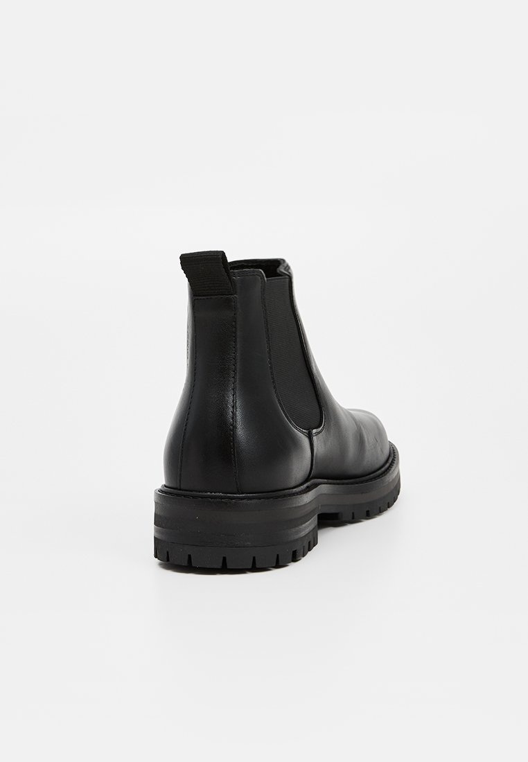 Schwarze Leder-Chelsea-Boots mit elastischem Seitenpanel, Zuglasche an der Ferse und dicker Profilsohle, von hinten auf weißem Hintergrund gezeigt.