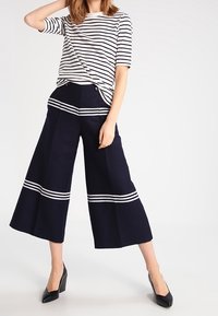 Žena oblečená v černých širokých zkrácených kalhotách s bílými pruhy a bílém tričku s černými vodorovnými pruhy, pózující s jednou rukou nahoře.