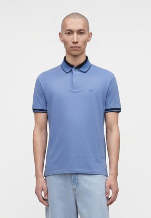 Homme à la tête rasée porte un polo bleu clair avec col et poignets foncés, ainsi qu’un jean bleu clair, debout devant un fond blanc uni.