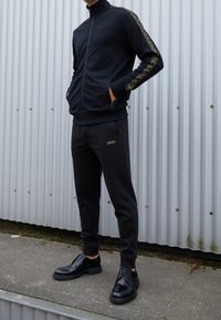 Chándal deportivo negro con chaqueta con cremallera y pantalones, con detalles de logotipo dorado. Tela suave y diseño ajustado. Zapatos negros con cordones.