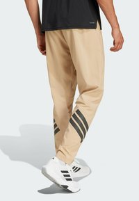 Beige sportbroek met zwarte gestreepte accenten, losse pasvorm en zachte stof, gecombineerd met witte sneakers met zwarte branding.