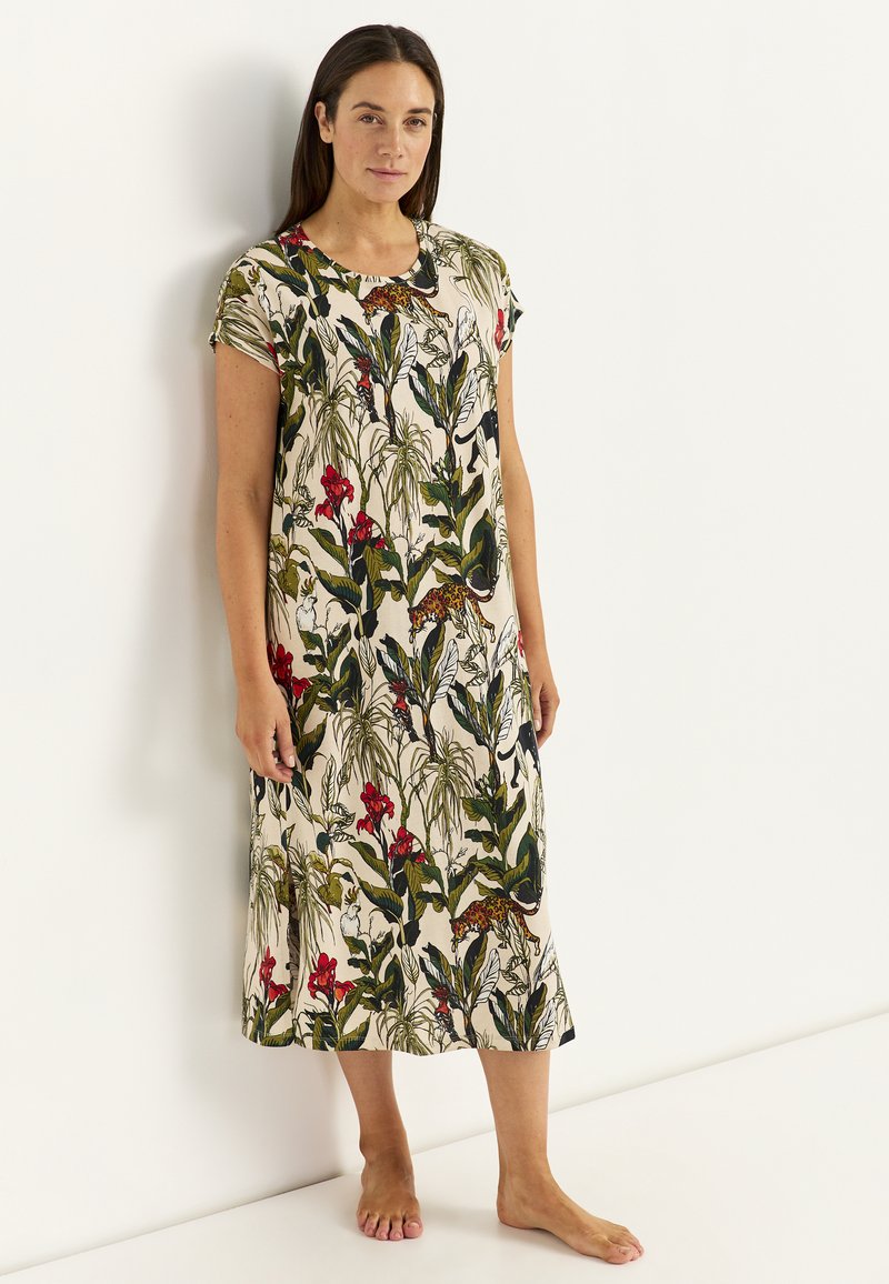 Frau, die barfuß an einer Wand steht, trägt ein knie langes cremefarbenes Kleid mit grünen Blättern, roten Blumen und Leopardenmuster.