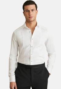 Cămașă albă cu un front texturat, guler clasic și butoane negre. Mânici lungi cu manșete simple, stilizată cu pantaloni negri.