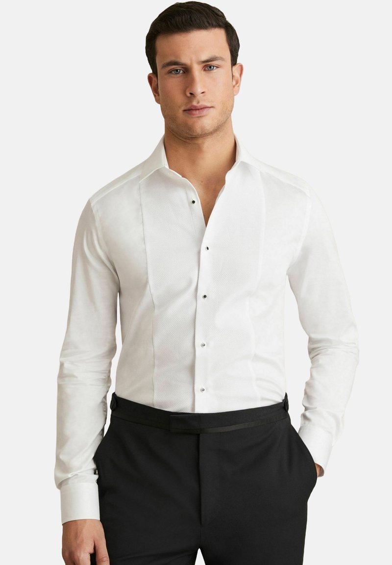 Cămașă albă cu un front texturat, guler clasic și butoane negre. Mânici lungi cu manșete simple, stilizată cu pantaloni negri.