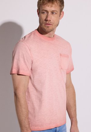 Uomo che indossa una maglietta rosa chiaro a maniche corte con taschino sul petto, guarda di lato su uno sfondo chiaro uniforme.