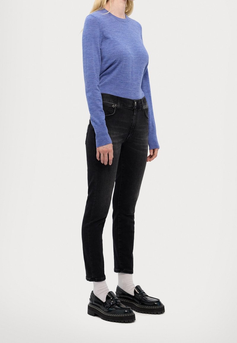 Femme debout, portant un haut bleu à manches longues, un jean slim noir, des chaussettes blanches et des mocassins noirs épais, sur un fond uni.