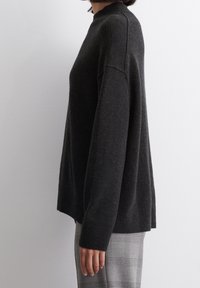Pull gris foncé avec un col montant, des épaules tombantes et des poignets côtelés, fabriqué en tissu doux. Associé à un pantalon à carreaux gris.