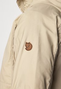 Veste technique beige au toucher lisse, avec un logo brodé marron d'un renard sur la manche gauche.