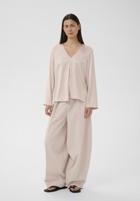 Femme aux longs cheveux noirs portant un haut beige ample à col en V, un pantalon à jambes larges et des sandales noires, debout devant un fond blanc uni.