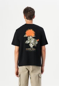 Schwarzes Baumwoll-T-Shirt mit einem bunten floralen Design auf der Rückseite, das orangefarbene Blumen und grüne Blätter zeigt. Minimalistischer Text unterhalb der Grafik.