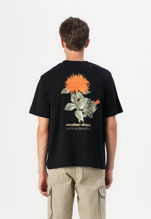 Sort bomulds-t-shirt med et farverigt blomsterdesign med orange blomster og grønne blade på ryggen. Minimalistisk tekst under grafikken.