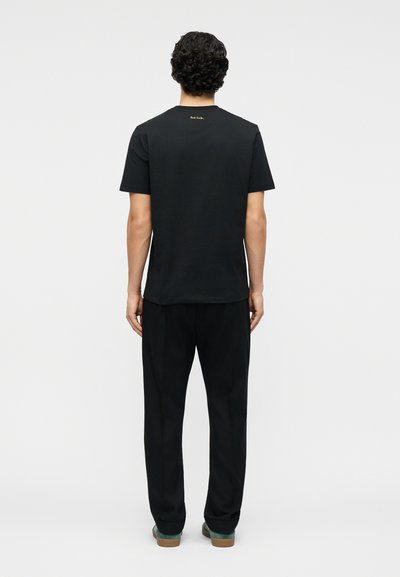 Άνδρας που στέκεται γυρισμένος με την πλάτη, φοράει απλό μαύρο κοντομάνικο t-shirt με μικρό λευκό κείμενο κοντά στον λαιμό και άνετο μαύρο παντελόνι.