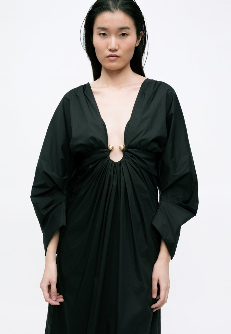 Abito nero, oversize, con una profonda scollatura a V e tessuto drappeggiato, dotato di un accento dorato al centro e maniche lunghe e soffici.