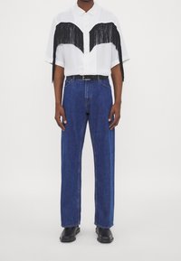 Camicia bianca a maniche corte con dettagli a frange nere, abbinata a jeans di denim blu larghi e stivali neri. Colletto standard e bottoni frontali.