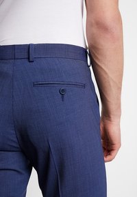 Marinblå dressbyxor i texturerat tyg, med en bakficka med knappstängning och bälteshängare.