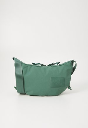 Grøn crossbody-taske i stof med justerbar rem og logo-patch foran, med snøre-detalje ved lynlåslukning.