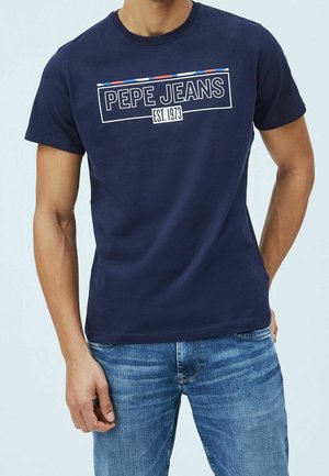 Homme portant un t-shirt bleu marine "PEPE JEANS EST. 1973" et un jean bleu, debout avec une main dans la poche, sur un fond clair.