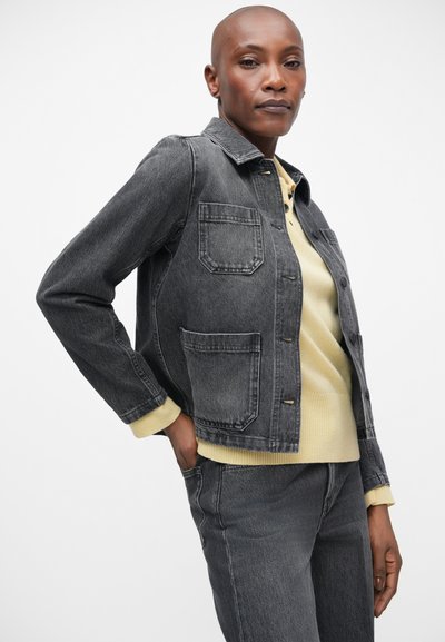 Veste en denim gris au design classique, avec deux poches avant, un fermoir à boutons et des poignets côtelés jaunes, superposée à un léger pull.