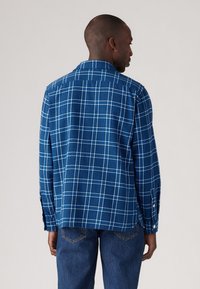 Chemise à carreaux bleus avec des lignes blanches, poignets à boutons, coupe décontractée et tissu texturé, portée sur un jean bleu, vue de dos.