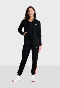 Felpa nera con zip sopra una maglietta nera, abbinata a pantaloni neri con una striscia arancione. Scarpe sportive bianche. Acconciatura riccia.