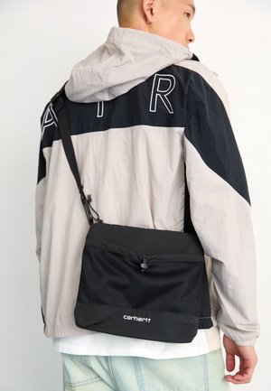 Personne portant une veste à capuche claire et noire avec le texte "AIR", et portant un sac bandoulière noir Carhartt sur l'épaule.