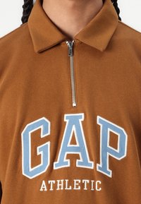 Brun zip-up sweatshirt med krage, utrustad med en stor ljusblå "GAP"-logotyp i fet stil och "ATHLETIC" under i vitt. Mjuk tygkänsla.