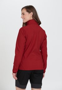 Röd softshell-jacka med hög krage, med en figursydd design och långa ärmar, kombinerad med svarta shorts. Slät textur och detaljering av sömmar.
