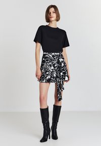 Femme portant un haut noir à manches courts, une jupe portefeuille florale noire et blanche, et des bottes à talons noirs jusqu'aux genoux, se tenant devant un fond uni.
