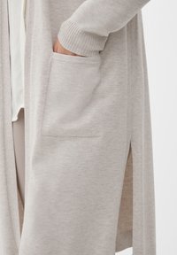 Ljusgrå cardigan i mjukt stickat tyg, med en slits på sidan, ribbade mudd och en framficka på nedre högra sidan.