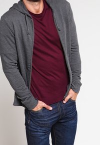 Grå zip-up hoodie med dragsko över en bordeaux t-shirt, tillsammans med mörkblå jeans. Slim fit, mjukt tyg och avslappnad design.
