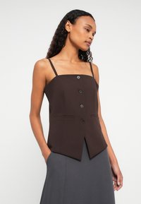 Sieviete ar gariem cirtainiem matiem, tērpusies bez piedurknēm brūnā pogājama blūzē un tumši pelēkās svārkās, pozē ar rokām kabatās uz balta fona.