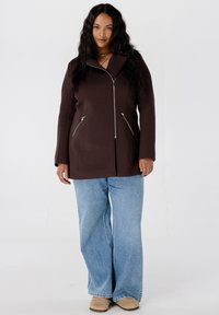 Bruine ritssluitingjas met een hoge kraag, twee zijzakken en zilveren hardware-accenten. Gecombineerd met lichtblauwe wijde jeans en beige schoenen.