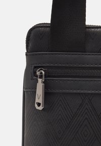 Svart handväska med ett geometriskt mönster, slät yta och en silverdragare med logotyp. Inkluderar ett robust tygband.