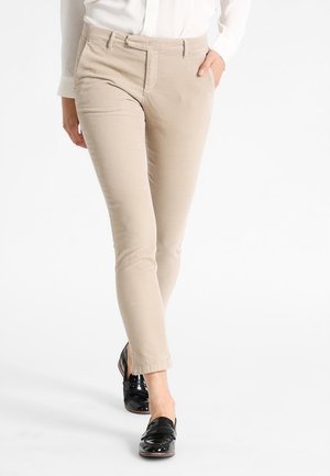 Person wearing beige slim-fit pants and black shiny loafers, walking with one hand in pocket against a white background.