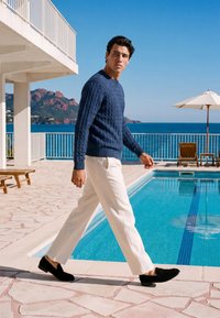 Homme portant un pull bleu et un pantalon blanc marchant près d'une piscine avec des chaises longues, un parasol et une vue sur l'océan en arrière-plan.