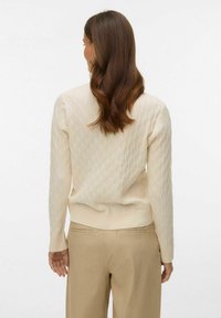 Kremowy sweter z dzianiny warkoczowej z prążkowanym ściągaczem na dole i mankietach, zestawiony z jasnobeżowymi spodniami. Gładka faktura, luźny krój.