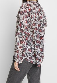 Blouse florale avec des manches volantées et un tissu léger et transparent. Présente des motifs floraux rouges et bleus sur un fond clair.